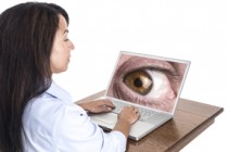 A young mixed race woman types on her laptop computer while she is being watched by a giant eye on the laptop screen.